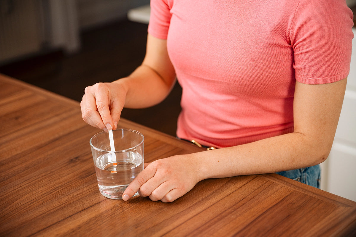 Person testing water hardness with a test strip in a glass of water on a wooden table.